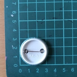 May include: A white button with a silver pin and the text "JM-32" printed on it.