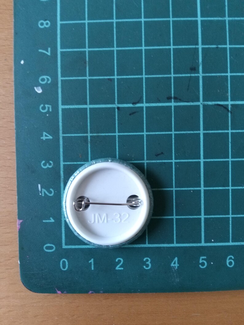 May include: A white button with a silver pin and the text "JM-32" printed on it.