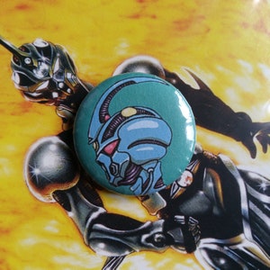 Puede incluir: Un botón azul y verde con un robot de dibujos animados con una cabeza grande y una sola antena. El robot tiene un cuerpo azul claro con detalles morados y ojos rojos.