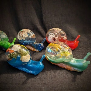 May include: Five colorful snail sculptures with intricate details. Each snail is made of resin and features a different color and design. The snails are arranged in a semi-circle on a dark background.