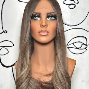 Puede incluir: Una peluca larga y lisa de color marr&oacute;n claro con sutiles reflejos rubios y raya en medio, expuesta sobre una cabeza de maniqu&iacute; con ojos verdes, pesta&ntilde;as largas y oscuras y labios rosados, con un borde frontal de encaje transparente.