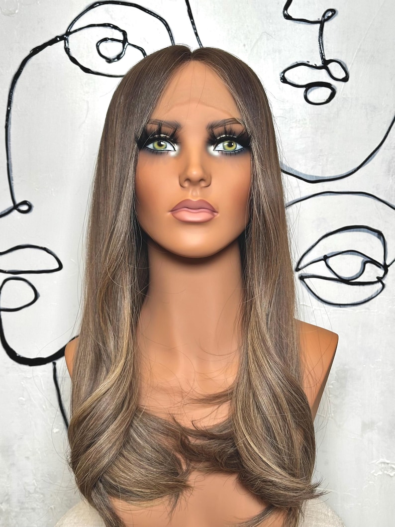 Puede incluir: Una peluca larga y lisa de color marr&oacute;n claro con sutiles reflejos rubios y raya en medio, expuesta sobre una cabeza de maniqu&iacute; con ojos verdes, pesta&ntilde;as largas y oscuras y labios rosados, con un borde frontal de encaje transparente.