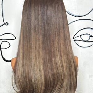 Puede incluir: Una peluca larga y lisa que presenta una paleta de colores casta&ntilde;os con tonos m&uacute;ltiples y reflejos rubios m&aacute;s claros. El cabello est&aacute; peinado con una raya en medio y cae por debajo de los hombros, mostrando una textura suave y un aspecto natural.