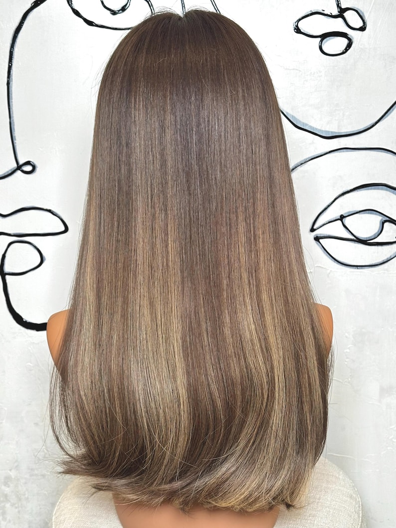 Puede incluir: Una peluca larga y lisa que presenta una paleta de colores casta&ntilde;os con tonos m&uacute;ltiples y reflejos rubios m&aacute;s claros. El cabello est&aacute; peinado con una raya en medio y cae por debajo de los hombros, mostrando una textura suave y un aspecto natural.