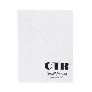May include: White fabric baby blanket with black text that reads "CTR Russell Monson November 18, 2023".