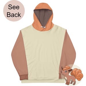 Puede incluir: Una sudadera con capucha de color crema con mangas marrones y una capucha marrón. La sudadera tiene un bolsillo grande en la parte delantera. La parte posterior de la sudadera presenta una imagen de dibujos animados de un Pokémon marrón y naranja.