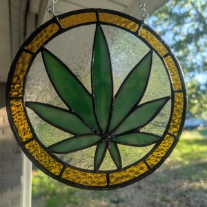 Puede incluir: Vidriera redonda con un diseño de hoja de cannabis verde. La hoja está centrada dentro de un círculo de vidrio transparente, rodeada por un borde amarillo texturizado y un marco negro. Una cadena plateada está adjunta para colgar.