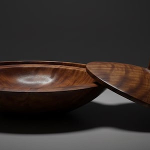 May include: A wooden bowl and lid, crafted from dark brown wood. The lid is partially open, displaying the bowl's interior. The wood grain is visible on both pieces. The bowl is round and has a smooth, polished finish. The bowl's diameter is approximately 20 cm.