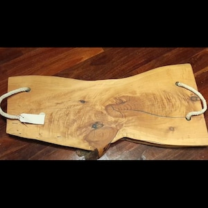 May include: A wooden serving tray with a natural, irregular shape. The tray is light brown with visible wood grain and knots. It has two white rope handles and a small tag attached. This rustic tray is suitable for serving food or as a decorative item.