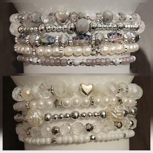 May include: Two stacks of beaded bracelets in shades of white, silver, and gray. The bracelets feature various beads, including pearls, faceted crystals, and metallic accents. Some bracelets have heart and star charms. The bracelets are displayed on a white cylindrical surface.