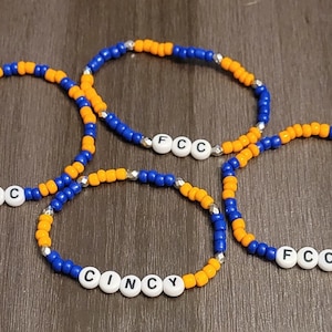 May include: Three beaded bracelets with alternating blue and orange beads. The bracelets spell out "FCC" and "CINCY" in white letters.