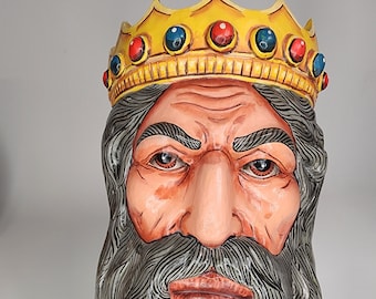 May include: A hand-carved mask of a king's face, with a yellow crown set with red and blue jewels. The face has a light skin tone, a long grey beard, and a stern expression. The mask is likely for costume or decorative purposes.