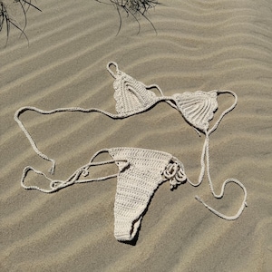 Puede incluir: Un conjunto de bikini blanco de ganchillo, compuesto por una parte superior y una inferior, sobre una playa de arena.