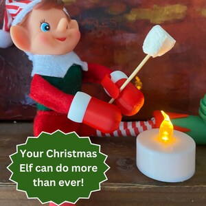 May include: A Christmas elf figurine wearing a red and green outfit with a matching hat, holding a wooden stick with a white marshmallow over a small, round, battery-operated white tea light candle with a glowing orange flame-shaped LED bulb.
