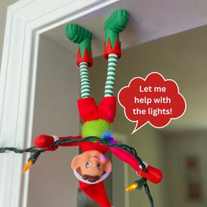 May include: A posable elf doll hanging upside down from a door frame, wearing a red and green outfit with striped leggings and green boots. The elf holds a string of orange Christmas lights and has a red speech bubble that says, Let me help with the lights!