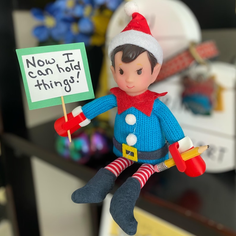 May include: A posable elf doll wearing a red and white hat, a blue knit sweater with white pom-poms, and red and white striped leggings with dark boots. The doll has red plastic hands holding a yellow pencil and a green sign reading Now I can hold things!