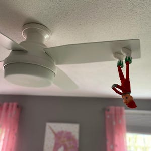 May include: A white ceiling fan with a small elf doll hanging upside down from one of the blades. The doll wears a red outfit with green accents and white shoes, with its feet attached to the underside of the flat white fan blade.
