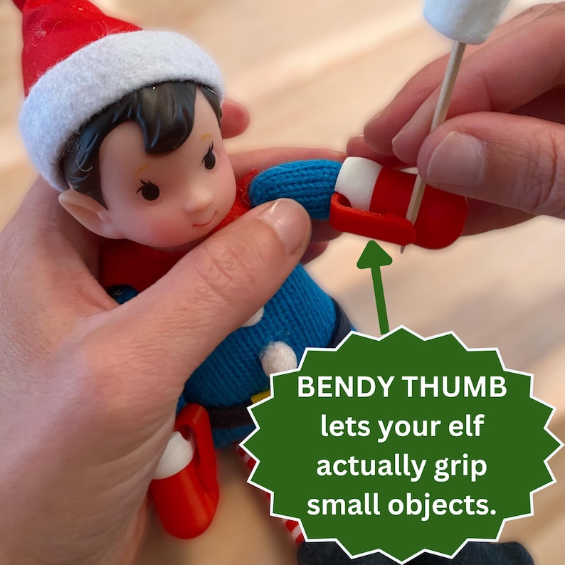 May include: A small elf doll wearing a red hat and blue sweater with white pom-poms. The doll features a red bendy thumb on its hand, which is gripping a wooden toothpick with a white marshmallow on top. Text reads: BENDY THUMB lets your elf actually grip small objects.