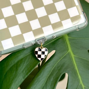 May include: A clear phone case with a green and white checkered pattern. A gold-toned charm in the shape of a heart with a black and white checkered design hangs from the case. The background features a large green leaf.