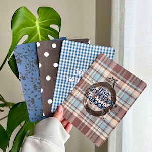 May include: A set of four rectangular cards with rounded corners. The designs include a blue card with dark leaf patterns, a brown card with white polka dots, a blue and white gingham card, and a brown plaid card with the text IT'S A GOOD DAY to read A BOOK.