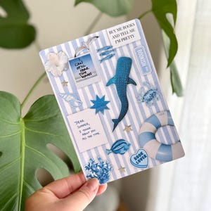 May include: A decorative card with a blue and white striped background, adorned with ocean-themed stickers. The card features a whale shark, starfish, and the text "BUY ME BOOKS AND TELL ME I'M PRETTY". Other text includes "CAN STILL HEAR THE WAVES" and "DEAR SUMMER, I THINK ABOUT YOU ALL THE TIME."