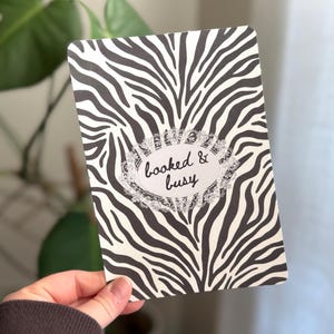 Op de afbeelding: Een rechthoekige kaart met een zwart-wit zebraprintpatroon. De kaart heeft de tekst "booked & busy" in een decoratief ovaal. De kaart wordt door een persoon vastgehouden.