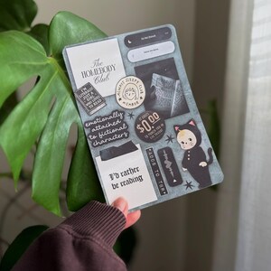 Könnte beinhalten: Eine dekorative Karte mit verschiedenen Aufklebern und Texten. Die Aufkleber enthalten Sätze wie "The Homebody Club", "I'd rather be reading" und "emotionally attached to fictional characters". Eine Katze im Kostüm ist ebenfalls abgebildet.
