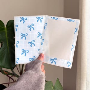 Pode incluir: Caderno aberto com um padrão de laço azul claro sobre um fundo de grade branco. As páginas internas estão em branco. O caderno é aberto por uma pessoa. O caderno é de cor azul claro e branco.
