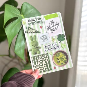Pode incluir: Uma folha de adesivos listrada em verde e branco com vários desenhos, incluindo um coelho de pelúcia, uma tigela de matcha e o texto "shh, I'm reading". Outros textos incluem "The Homebody Club" e "Lucky Charm".