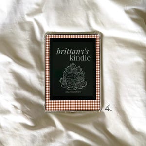 Può includere: Lettore di ebook Kindle in una custodia trasparente con bordo a quadretti marrone e bianco. Lo schermo mostra "brittany's kindle" sopra una pila di pancake con fragole. Il testo "my personal library" è sotto.