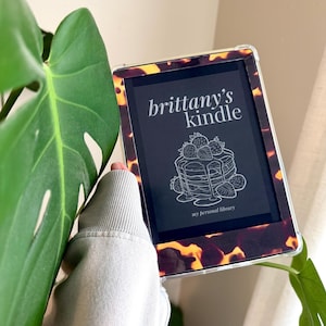 May include: An e-reader with a tortoiseshell-patterned case. The screen displays "brittany's kindle" above a pancake illustration with strawberries and the text "my personal library". The device is held near a large green leaf.