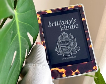 May include: An e-reader with a tortoiseshell-patterned case. The screen displays "brittany's kindle" above a pancake illustration with strawberries and the text "my personal library". The device is held near a large green leaf.