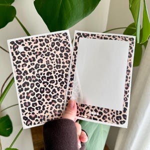 Op de afbeelding: Twee rechthoekige stickers met een luipaardprint in tinten beige, bruin en zwart. Eén sticker heeft een gat aan de bovenkant en de andere heeft een helder venster. De stickers worden voor een groene plant gehouden.