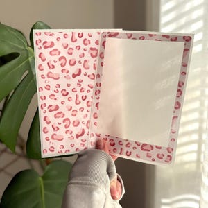 May include: A rectangular greeting card featuring a pink and white leopard print pattern on the exterior and a plain white interior panel. The card is open, displaying both the patterned outer cover and the blank white writing surface inside.
