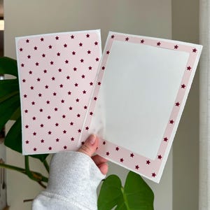 Peut inclure: Deux cartes de vœux sont présentées, une ouverte et une fermée. La carte fermée a un fond rose avec un motif d'étoiles rouge foncé. La carte ouverte a un intérieur blanc avec une bordure rose et des étoiles rouge foncé.