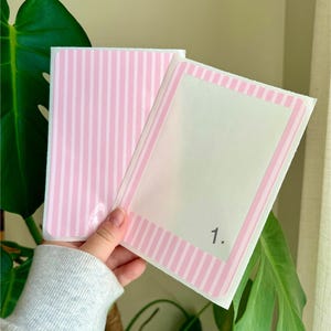 May include: Two pink and white striped cards, one with a blank white space and the number "1." in the corner. The other card has a pink and white striped pattern. The cards are held in front of a green plant.