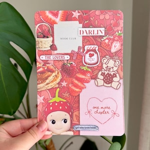 May include: A pink and red book club card with strawberry illustrations and the text "DARLIN" and "BOOK CLUB". The card features a strawberry-themed design with a teddy bear, a baby doll, and the text "one more chapter".