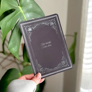 Op de afbeelding: Een donkergrijze rechthoekige kaart met zilveren details rond de randen. De kaart bevat de tekst "But daddy I love him" in sierlijk schrift. De kaart wordt voor een groene plant gehouden.