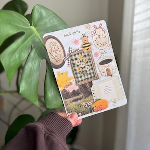 Peut inclure: Un journal décoratif avec le texte "book girlie" et divers autocollants, dont une abeille, des fleurs et une tasse de café. Le journal est tenu devant une grande feuille verte.