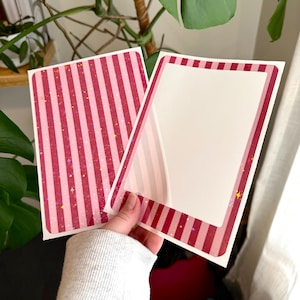 Puede incluir: Dos pegatinas rectangulares con un patrón de rayas rosas y rojas. Una pegatina tiene un diseño de rayas sólidas, mientras que la otra tiene un interior blanco con un borde a rayas. Ambas pegatinas tienen detalles de purpurina.
