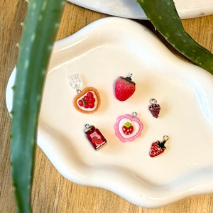May include: A selection of strawberry-themed charms. The collection includes a strawberry tart, a jar of jam, and several strawberry-shaped pendants. The charms are red, pink, and white, with silver-coloured metal findings.