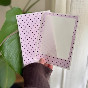 May include: A rectangular card featuring a light pink background with a pattern of small black polka dots. The card has a central white rectangular area framed by a border of the same pink and black polka dot pattern, creating a frame effect.