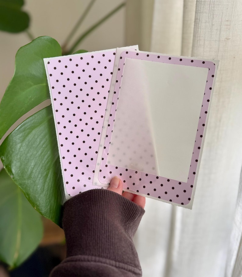 May include: A rectangular card featuring a light pink background with a pattern of small black polka dots. The card has a central white rectangular area framed by a border of the same pink and black polka dot pattern, creating a frame effect.