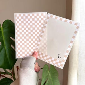 Op de afbeelding: Twee doorzichtige plastic hoezen met een beige en wit geblokt patroon. Eén hoes is volledig bedrukt, terwijl de andere een geblokte rand heeft en het nummer "1." in zwart is gedrukt. De hoezen worden tegen een neutrale achtergrond gehouden.