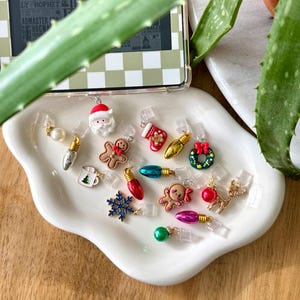 Puede incluir: Una colección de abalorios con temática navideña, que incluye un Papá Noel, un hombre de jengibre, luces navideñas y un copo de nieve. Los abalorios son de varios colores y se muestran en un plato blanco con bordes ondulados.