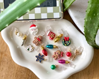 May include: A collection of Christmas-themed charms, including a Santa Claus, gingerbread man, Christmas lights, and a snowflake. The charms are in various colors and are displayed on a white, wavy-edged dish.