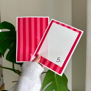May include: Two rectangular cards. One card has vertical stripes in shades of red and pink. The other card has a white interior with a red and pink border and the number 5 in the bottom right corner. The cards are held in front of a green plant.