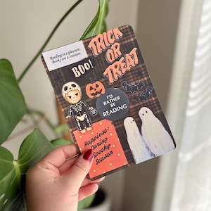 May include: A rectangular card with a brown plaid background featuring Halloween-themed stickers including a skeleton, a pumpkin, bats, and two ghosts. Text reads: Reading is a pleasure. Books are a treasure. TRICK OR TREAT, BOO!, I'D RATHER BE READING, and Happiest: During Spooky Season.