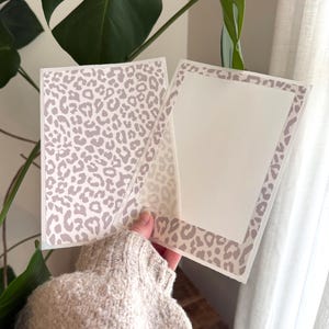 May include: A rectangular greeting card featuring a light beige and white leopard print pattern. The card is open, revealing a solid white interior panel framed by the same leopard print border on the edges, providing a clean space for writing.