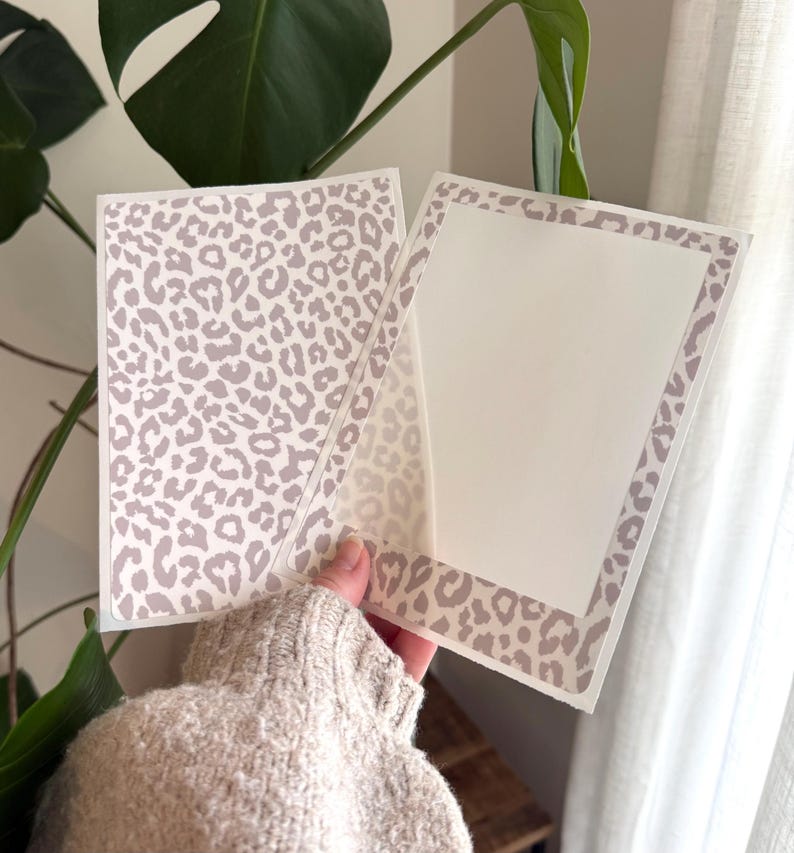 May include: A rectangular greeting card featuring a light beige and white leopard print pattern. The card is open, revealing a solid white interior panel framed by the same leopard print border on the edges, providing a clean space for writing.